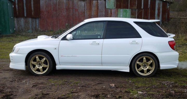 Pistonheads - The image shows a small white hatchback car parked on a patch of grass next to a rusty red-brown building with a corrugated metal roof. The car appears to be in good condition, with golden-silver two-tone rims on its wheels. The photo's composition has the car positioned on the right, and the building is on the left, creating an interesting contrast of the shiny vehicle against the aged structure. There's a hint of smoke coming from the car's rear, suggesting recent or imminent movement.