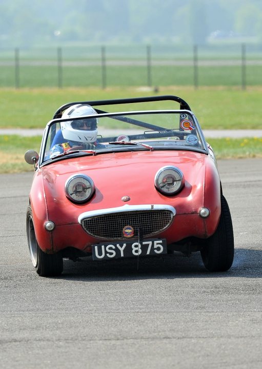 COOL CLASSIC CAR SPOTTERS POST!!! - Page 384 - Classic Cars and Yesterday's Heroes - PistonHeads - The image shows a vintage red convertible car with a smiling grill, resembling a classic model, likely an early Subaru model, based on the distinctive grille design. The car is in motion with its top down, and the driver is visible wearing a white helmet and pilot gear. The setting appears to be an outdoor track with a grassy area to the side. The overall style of the image suggests it is a photograph of a car race or event.