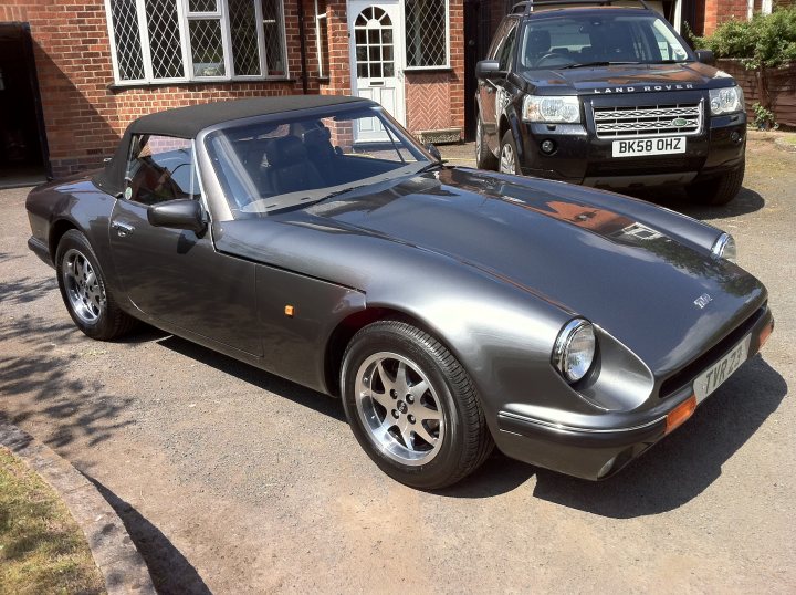 TVR23 V8S Body Off Restoration - Page 1 - S Series - PistonHeads - The image shows a black sports car parked in a driveway. The vehicle appears to be a Porsche 944 Slant Nose, characterized by its distinctive aerodynamic design, sloped front end, and large, headlight style fog lights. Additionally, it features a set of chrome wheels. In the background, there is a brick house with a white front door. To the right of the car, there is another vehicle, likely an SUV. There are no people visible in the image. The setting appears to be a residential area.