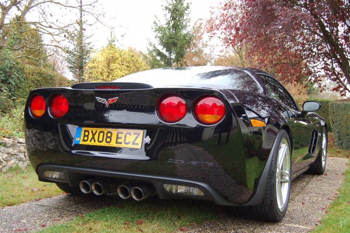 Pistonheads - The image depicts a black sports car parked on a gravel driveway with grass on either side. The car has a glossy finish and features the Corvette emblem, suggesting it's from the General Motors brand. It has a clean and sleek design, with visible taillights and exhaust pipes. The license plate reads "BX08 ECR". The surrounding area includes trees and autumn foliage, indicating it could be late summer or early autumn.