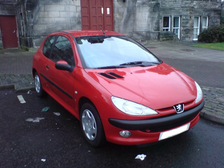 Up yours Gordon ! - Page 1 - Readers' Cars - PistonHeads - The image depicts a red hatchback car parked in a lot. The car has a distinctive front grille with a logo, which is indicative of the automaker. The vehicle's body shows a glossy finish, reflecting some of the surrounding environment. There are buildings in the background, suggesting an urban or suburban setting. A greenish hue to the upper part of the image gives a serene atmosphere, contrasting with the car's bright red color.