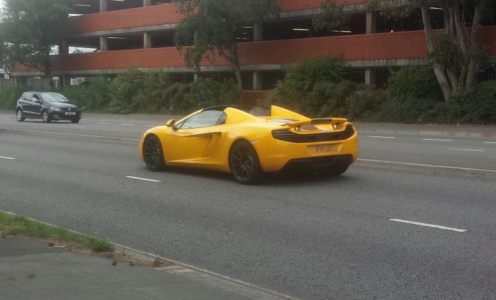 Spotted In South Wales (Vol 3) - Page 213 - South Wales - PistonHeads - The image shows a bright yellow sports car driving down a street. The car is distinctive with its two doors and prominent air intakes at the rear of the hood. It appears to be a high-performance model, possibly an Audi R8 or a similar type of vehicle. The car is the central focus of the image, moving from left to right across the frame. The street itself seems to be a residential or suburban area with parked cars and what looks like a parking lot in the background.