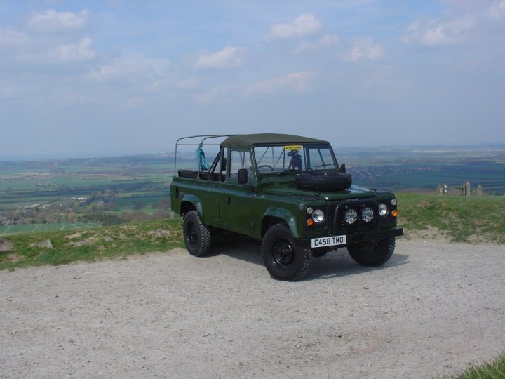 Pistonheads Roof Defender - Image depicts a four-wheeled vehicle, commonly known as a truck or jeep, parked on a gravel surface. The vehicle is green with a horizontal bar at its rear declaring "450 TMW". The landscape in the background appears to be a countryside area with rolling hills and open fields under a partly-cloudy sky. The vehicle's spare tire is resting on the back of the vehicle.