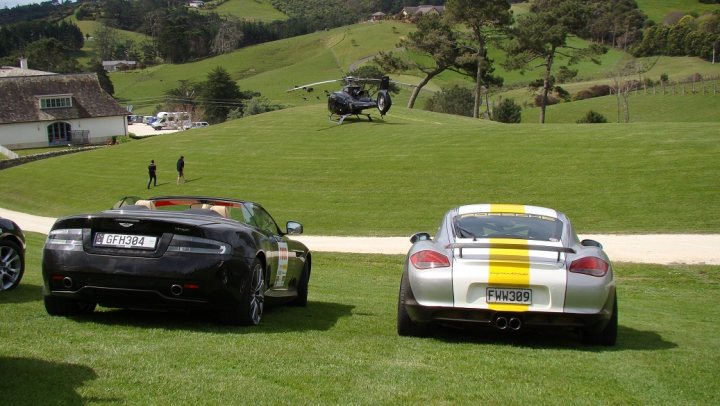 Targa 2013 - Page 1 - New Zealand - PistonHeads - The image captures a serene outdoor setting where several cars are parked on a grassy slope. Among the cars, a black Bentley, a black sports car, and a silver convertible stand out. A yellow helicopter is prominently positioned above the scene, seemingly preparing for takeoff. The setting includes a white house nestled among the green grass and hills. The overall atmosphere of the image exudes luxury and elegance.