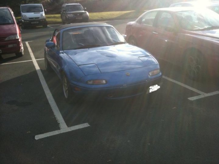 £3k, too steep? plus introduction - Page 1 - Mazda MX5/Eunos/Miata - PistonHeads - In this photograph, a blue coupe sports car is parked in an otherwise deserted parking lot. The car's vibrant color stands out against the muted tones of the surrounding environment. The parking lot itself is empty, save for a few other parked vehicles scattered in the distance, their more neutral colors serving as complements to the car's striking blue. The photo seems to have been taken on a sunny day, as indicated by the radiant sunlight illuminating the scene.