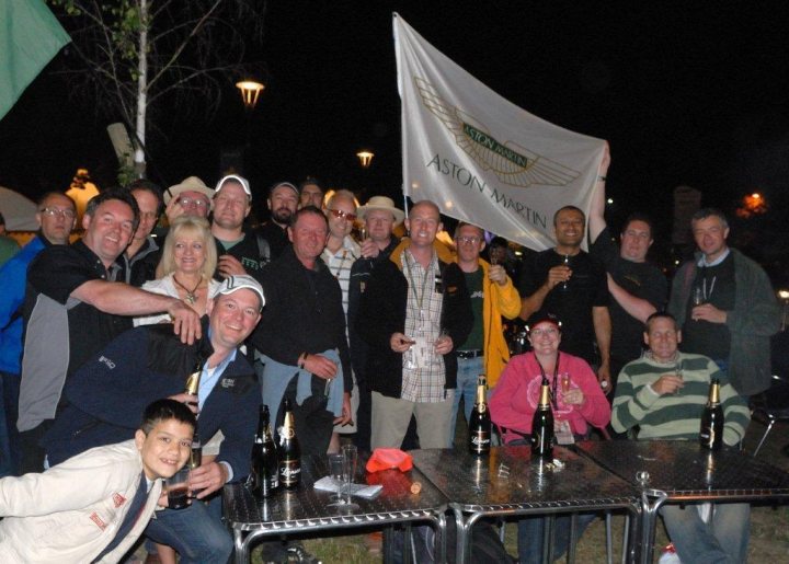 Le Mans - Page 1 - Aston Martin - PistonHeads - The image captures a lively scene of a group of people celebrating an event. They are standing around tables and holding a large white flag with an eagle emblem, which suggests a festive atmosphere. The tables are adorned with champagne bottles, glasses, and other party essentials. The mood is joyful, as evidenced by the smiles on the attendees' faces. The event appears to be taking place at night, as indicated by the darkness in the background.