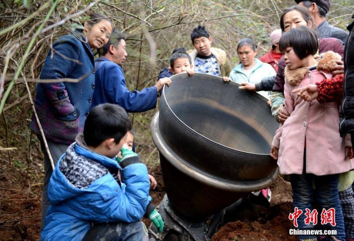 A group of people standing next to each other - Pistonheads - The image shows a group of people, primarily Asian, gathered around a large metal object in an outdoor setting, which appears to be muddy. The object in question resembles one that might have been used in a makeshift water purification setup. The group seems to be inspecting or discussing this item. The expressions on their faces suggest a mix of curiosity and concern. The mood of the image is somewhat puzzled, with the people trying to understand the object's purpose.