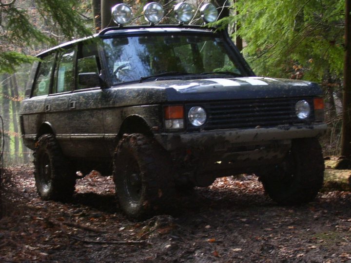 Classic Pistonheads Range Rover Beast - The image features a sturdy, off-road vehicle in a wooded area. The vehicle, which appears to be a SUV or a jeep, is covered in a substantial layer of mud, indicating it has likely been traveling off paved surfaces. The vehicle is equipped with a roof rack, and there are four headlamps visible. The surrounding environment is forested and the ground is rocky and muddy, suggesting a rugged outdoor setting.