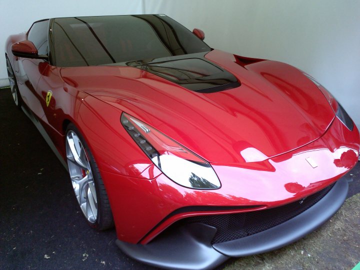 La Ferrari at FOS - Page 1 - Goodwood Events - PistonHeads - This image features a vibrant, high-performance red sports car parked indoors with a minimalist background. The vehicle displays a shiney exterior with a striking red paint job on its body and black grilles accenting the front and back of the car. It has aerodynamic wheel arches and a low-profile design, suggesting it's a high-performance model with enhanced efficiency and speed capabilities. The rims of the wheels appear to be highly polished, reflecting the car's overall sleek look. The car is positioned facing us, allowing us to see its full profile and the gleaming design elements.