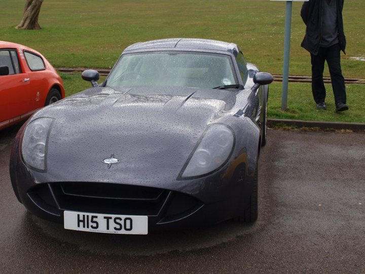 Marcos Display - Clevedon 14/04/2013  - Page 1 - South West - PistonHeads - The image features a gray sports car parked on the side of a road. A sign stands prominently beside the car. The car and the sign are in sharp contrast to a grassy field that occupies the background. The sky above is overcast, suggesting cloudy weather. The image is taken from a distance, slightly askew, giving a clear view of the front of the car.