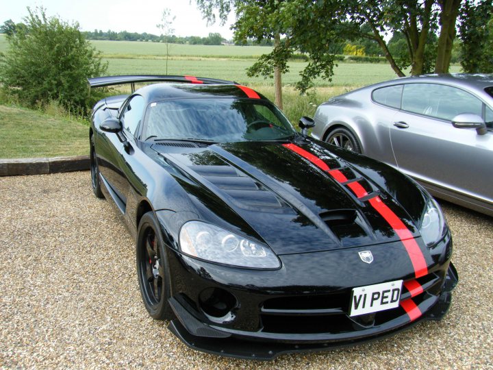 Breakfast Meeting, The Hare, Roxwell 4th June - Page 1 - Kent & Essex - PistonHeads - The image showcases a striking black sports car parked on a gravel surface. The car features a prominent rear spoiler and a seam running down the hood and roof. Red accents add a pop of color to the vehicle, with the taillights also displaying red elements. Adjacent to the sports car is a silver car, parked on the same gravel drive. The vehicles are positioned in front of a lush green field and a large tree, creating a contrast between the man-made precision of the cars and the natural environment.