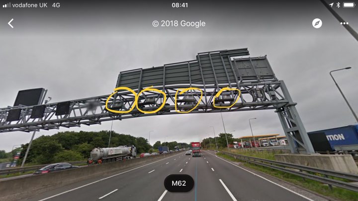 Are these speed cameras? - Page 1 - Speed, Plod & the Law - PistonHeads - The image shows a bustling highway scene viewed from a car's navigation system displayed on a smartphone. An overhead digital signboard indicates that there are eleven miles (presumably miles) to the destination M25. In the background, the highway is lined with lush greenery, and various vehicles, including a prominent tanker truck, are visible on the road.