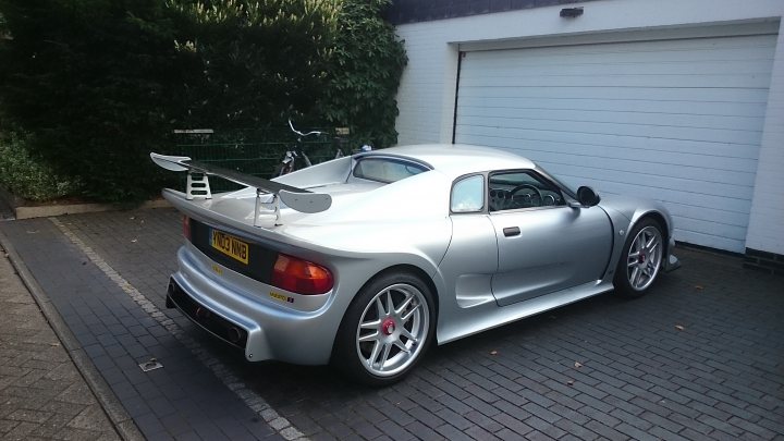 A white car parked in front of a building - Pistonheads - The image shows a silver sports car parked outdoors next to a garage. The car has an extended wing on the rear, which is characteristic of certain sports car models designed for aerodynamic downforce during high-speed driving. The wing has an early design reminiscent of classic or retro racing cars. The garage has a side door, and the ground appears to be a brick driveway. In the background, there's a hint of greenery, suggesting the area might be part of a residential property.