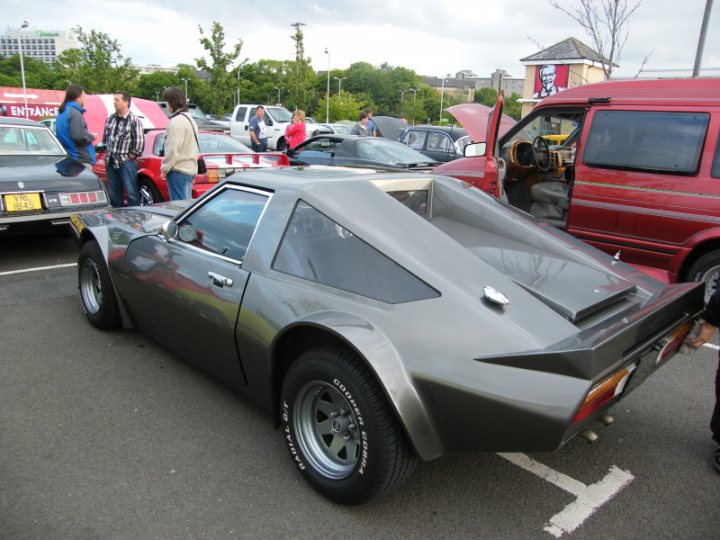 Cruisemeet Pistonheads Edinburgh - The image depicts an outdoor scene of a car show, with a focus on a unique vehicle. This vehicle appears to have a design that is open at the rear, revealing a configuration of exhausts and what might be a roll bar, indicative of a sports or racing application. The car is next to a red van with the doors open, possibly allowing spectators closer examination. 

In the background, there are various other vehicles and people, giving a relaxed and casual atmosphere to the event. The presence of trees and other structures suggests that the event is taking place in a spacious, outdoor area, possibly a large parking lot or a dedicated car show venue. Overall, the image conveys the excitement and interest of a car show, with enthusiasts gathered to appreciate and discuss the vehicles on display.