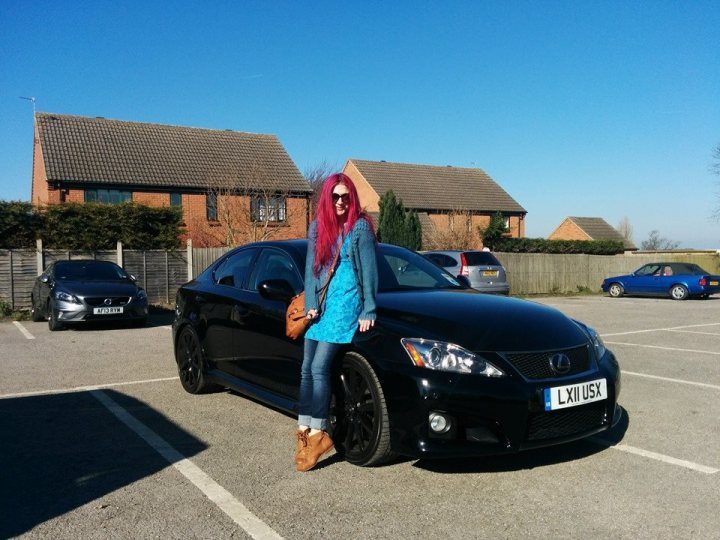 New IS F  - Page 1 - Jap Chat - PistonHeads - The image depicts a young woman standing by a black 4-door car. She has long, vibrant red hair and is casually dressed in a blue top and jeans, along with brown boots. She is smiling and appears to be gazing beyond the camera into the distance. The car is parked in a parking lot, and in the background, there are other vehicles. The sky is clear and blue, suggesting it might be a sunny day. The architecture of the buildings behind the cars has a brick facade and pointed roofs, giving the scene a small-town or suburban feel.