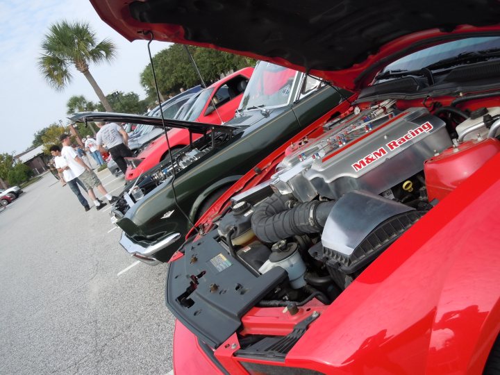 Cars & Coffee Charleston, SC - Page 1 - USA & Canada - PistonHeads - The image shows a close-up view of an engine bay in a red sports car. The hood of the car is open, revealing a complex network of tubes, wires, and mechanical components. The make of the engine can be seen, with the name "MAZDA" prominently displayed on various parts. The car is parked on a lot, surrounded by other vehicles, and there are a few people standing nearby, suggesting a possible auto show or racing event.