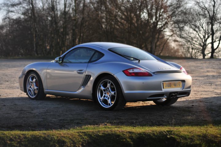 Boxster & Cayman Picture Thread - Page 10 - Boxster/Cayman - PistonHeads - The image shows a silver Porsche sports car parked on what appears to be a gravel surface or a dirt road with sparse grass, suggesting an outdoor, natural setting. The vehicle is captured from a slightly elevated angle, highlighting its sleek design and alloy wheels. The sunlight is diffused, casting soft shadows and a gentle gradient across the car. In the background, there is a line of trees and what might be a hill or a slope, adding a tranquil and picturesque atmosphere to the scene.