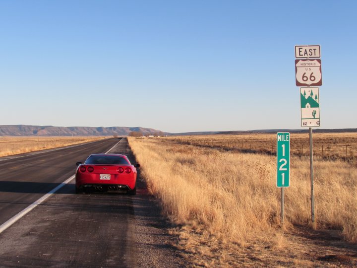 The £7700 Corvette C6 - Page 1 - Readers' Cars - PistonHeads - The image depicts a rustic highway scene under a clear blue sky. A red sports car is parked on the edge of a paved road, shaded by a large tumbleweed plant on the right side of the road. The car is prominently featured in the foreground, indicating a sense of adventure or travel. East is the direction indicated by the highway signs ahead, and there's an icon signifying cattle or livestock crossing Route 66. The sky appears to be sunny and clear, suggesting it's daytime.