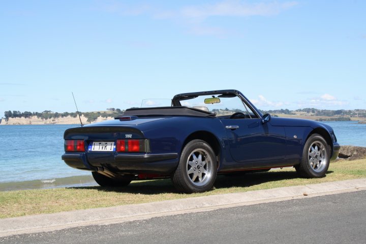 Roof down pictures - Page 1 - S Series - PistonHeads - The image displays a classic blue convertible sports car parked on the side of a road by the sea coast. The setting is during daylight, with the car's top down, offering an unobstructed view of the interior. The vehicle has a distinctive design, featuring a large rear spoiler, slender headlights, and a curved front fender. The rustic beach and the expansive blue ocean provide a contrasting backdrop to the sleek, modern vehicle.