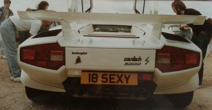 Countach  - Page 9 - Lamborghini Classics - PistonHeads - The image is a photograph that captures a close-up view of the rear of a sports car with a distinctive design. The car is identifiable by its white body, the spoiler on the hood, and the unique taillight arrangement, featuring red and orange lights. There is text visible on the upper part of the car, including the word "Audi," and the license plate reads "18 SEXY." In the background, there are at least three people who appear to be attendees at an auto show or event. The setting looks like an open area or parking lot with large vehicles nearby. There's a sense of motion and activity associated with the occasion.