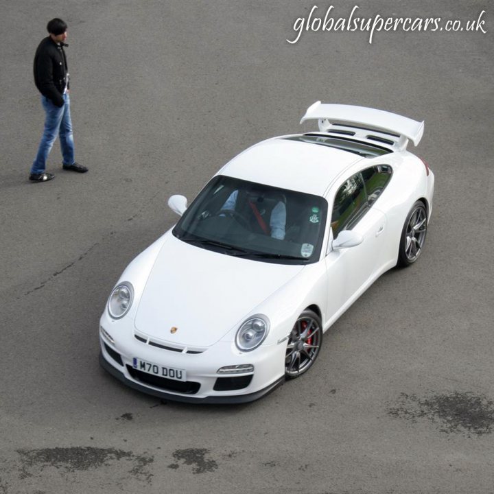 Snaps Service Silverstone Sunday Pistonheads Supply - The image features a white Porsche roadster parked on what appears to be a street or a circuit. In the foreground, a person is standing on the tarmac, seemingly observing the car. The Porsche has a sleek design, and there are visible tires and a distinctive rear spoiler. The background is somewhat blurred, but there is an empty lot or pavement surrounding the car.