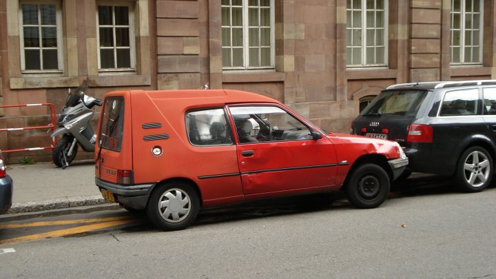 A yellow taxi cab parked on the side of a road - Pistonheads - The image shows a small red car parked in front of a large black SUV. The car is smaller and has a distinctive red paint. It is positioned on a street, and the larger vehicle is parallel-parked behind it. There is a gray motorcycle parked behind the car. The scene takes place in front of a building, which appears to be an apartment or residential building, as suggested by the windows and the construction of the facade. The buildings' architecture is traditional, with brickwork and multiple windows visible in the background. The perspective is slightly slanted, with the camera likely on a tripod.