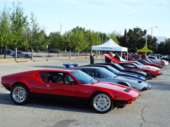 My 1972 De Tomaso Pantera - Page 2 - Readers' Cars - PistonHeads - The image shows a parking lot filled with vintage cars, prominently featuring a red DeLorean among other classic models. These vehicles are parked in rows under the open sky, suggesting an outdoor gathering or car show. A tent is set up near the cars, indicating an event or sale. There are people visible in the background, likely attendees of the event. The setting appears to be a sunny day with clear visibility.