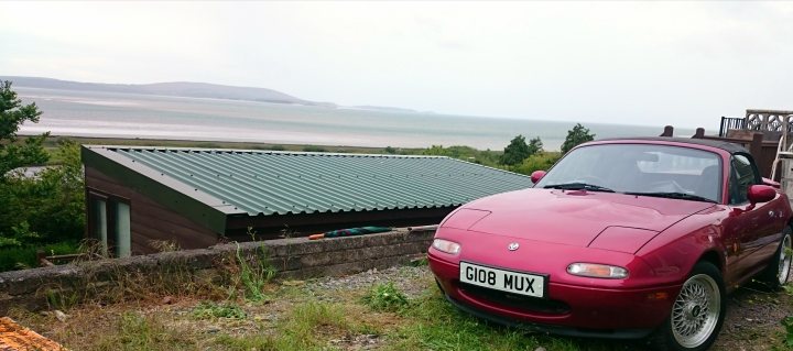 Pistonheads - The image shows a red sports car parked on a grassy area which appears to be a driveway or a small hillside. Beyond the car, there is a picturesque view of a large body of water, likely an ocean or a sea, with mountains or hills visible in the distance under a partly cloudy sky. There is a small, green hut or shed with a tiled roof near the car, suggesting a recreational or rural setting. The vehicle has a UK-style license plate. The scene suggests a peaceful, possibly remote, location with scenic views. The car is parked facing the water, indicating someone might have parked it to enjoy the view.