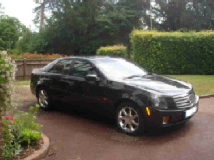 Cts Sale Pistonheads Cadillac - The image features a small, black luxury car parked in a driveway. The car is positioned at an angle, showing the front and side profile. The surrounding environment includes a verdant hedge, a bush, and a glimpse of a house in the background. The setting conveys a tranquil suburban scene.