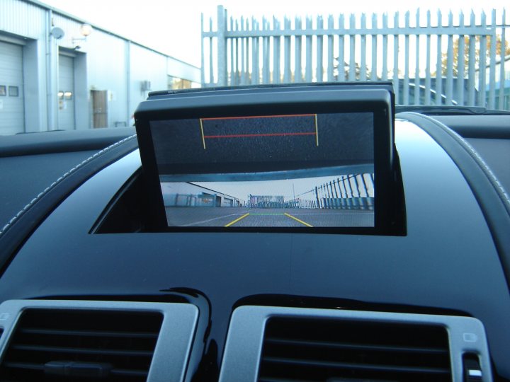 Pistonheads - The image provides a view from the passenger side of a vehicle, focusing on a rear-view mirror with a small, digital display embedded in the center. The display shows a road view with fencing along the side of the road and a blurred building in the distance, suggesting a city or suburban environment. The vehicle's interior is glossy and sleek, characterized by the use of a dark color scheme. The outdoor setting appears to be a one-story building in the background. The lighting suggests it is daytime with clear skies.