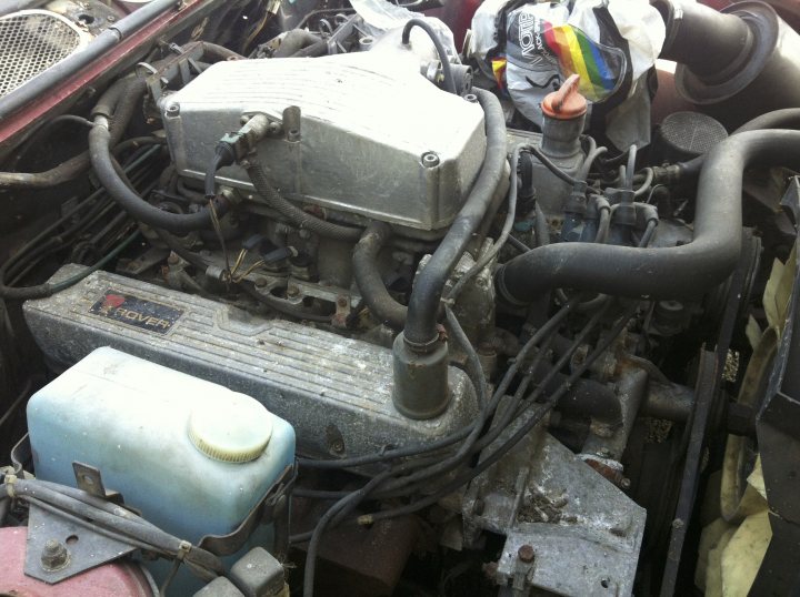 Pictures of my "NEW" SD1 Vitesse - Page 1 - Rover - PistonHeads - The image provides a close-up view of an engine bay in a vehicle. The engine is old with visible dirt and rust, suggesting it's not been maintained in a while. A water pump and a radiator are among the mechanical components visible in the picture. There is a capacity gauge on the front of the engine, which is typically used to estimate the motor oil capacity needed in a vehicle. Interestingly, there's also a rainbow-colored windshield wiper in the image, adding a splash of color to the otherwise monochromatic scene. The address visible in the image is of this car's owner, who could probably use a hand with some car maintenance suggestions!