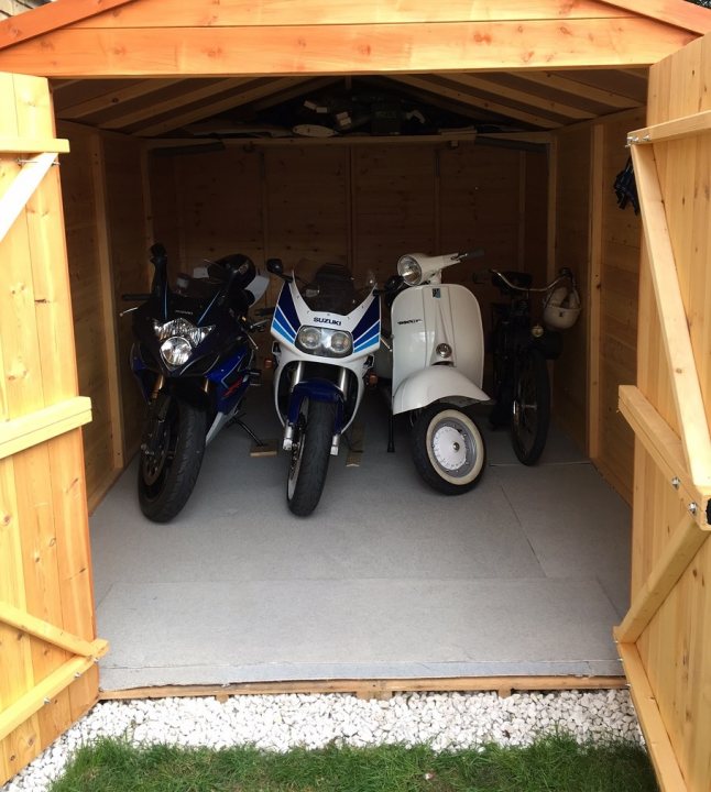 Bike in an 8x6 shed!? - Page 1 - Biker Banter - PistonHeads - The image shows a distinctive scene featuring three motorcycles parked inside a wooden garage or shed. The most prominent motorcycle is the one in the middle, which is white and blue with the word "Aprilia" visible on the front of the fairing. To the left, partially obscured, is another motorcycle with a primarily black color, and to the right, a third motorcycle is parked. The ground of the garage is meticulously covered with grey pebbles, creating a picturesque contrast with the warmth of the wooden structure. The overall setting suggests a motorcycle enthusiast's haven, with the motorcycles well cared for and kept in a sheltered location.