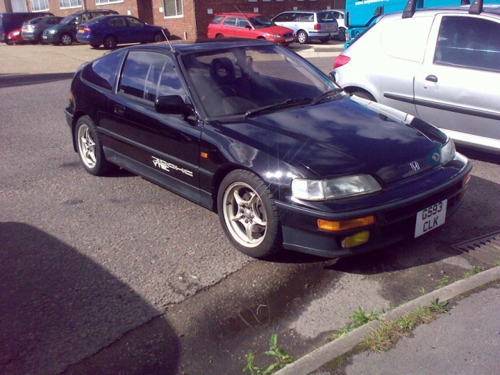 Pistonheads Member Sir Crx - The image shows a black Honda Civic Type R parked on the side of a road. The car is a sports model, identifiable by the aggressive body kit and wider wheel arches. It features a large rear spoiler and black side mirrors with repeaters, all of which are typical for Type R models. These modifications usually improve aerodynamic downforce, often for better handling and speed on the track. Behind the car, there are various vehicles and buildings, suggesting a busy urban environment. The car's license plate is partially visible with the letters 'G53'. The image is taken in a narrative style, capturing a typical day with vehicles parked on the side of a road.