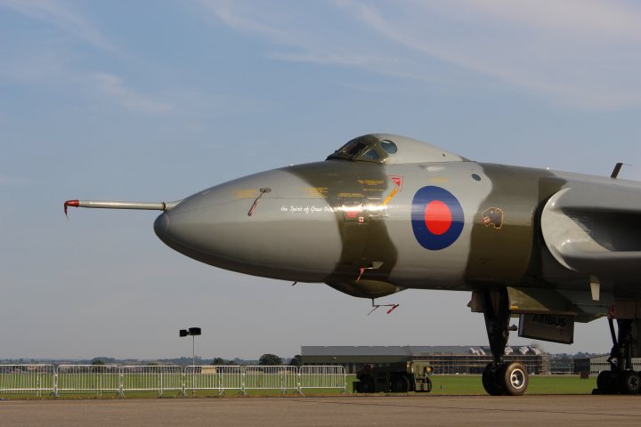 A small plane is parked on a runway - Pistonheads - The image features a military jet parked on a runway. The jet is a prominent silver aircraft with markings and red tracking spots visible on its outer surface. The cockpit features a black canopy that provides protection to the pilot. The aircraft is stationed in a clear blue sky, possibly in the foreground of an air force base or similar setting. The runway beneath the jet is expansive and flat, leading to green grass and distant buildings and a factory.