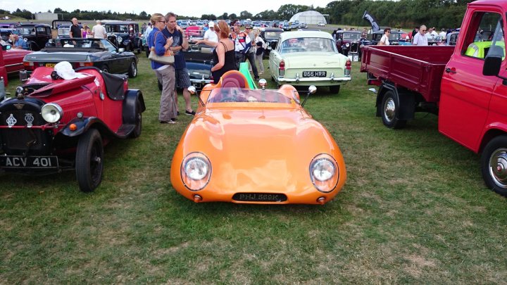 Drive out HalfWay House to North Weald sunday 3rd Aug  - Page 1 - Kent & Essex - PistonHeads - In the heart of a densely green field, a group of people stand admiring a row of vintage and classic cars. A woman, standing near a vibrant yellow car, seems in conversation with a man beside her. The vehicles, varying from shades of orange, yellow, and red, are parked in two rows on the grass. The open field extends into the distance, lined with trees that provide a natural backdrop to the event. The scene exudes a sense of leisure and nostalgia.