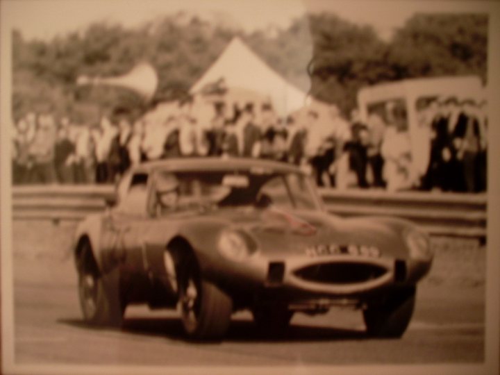 Raceing Wherewhen Etype Pistonheads - The image is a vintage black and white photograph featuring a driver in a classic race car on a racetrack. The car, adorned with an elaborate design, has sharp tail fins and is driving on what appears to be the end of the day, as suggested by the shadow on the track. The backdrop is a crowd of spectators watching from the grandstands, punctuated by a blurred solitary tall building, likely a part of the trackside infrastructure. The atmosphere is tinged with nostalgia, capturing a moment of automotive history.