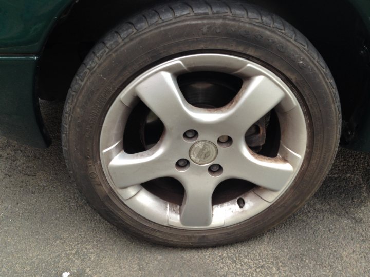 are these original Mk1 wheels on my mx5? - Page 1 - Mazda MX5/Eunos/Miata - PistonHeads - The image shows a black round tire with a silver rim and hubcap, positioned next to a green car. The tire is tilted at an angle suggesting movement or just parked like that. The silver rim has a visible center piece and five bolts that secure it on the hubcap and the car's wheel well. The ground beneath the tire appears to be grey with a few specks of what might be dirt or gravel.