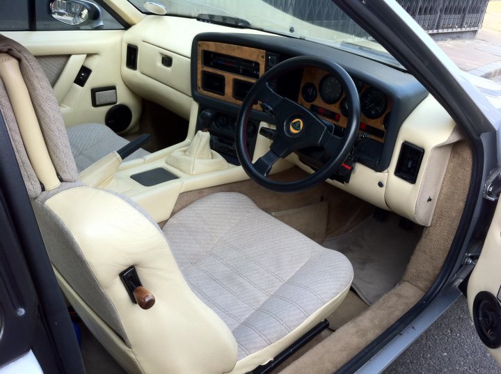 The Lotus is dead, long live the Lotus! - Page 6 - Classic Cars and Yesterday's Heroes - PistonHeads - This is an image of the interior of a vintage car, showing the steering wheel, gear shift, and part of the passenger seat. The car appears to be a convertible with two doors. The seat is white and seems to be upholstered in a vinyl or leather-like material. The steering wheel is black with a leather material and the gear shift is also black. The car has a dashboard that takes up much of the area and features a centrally placed glove box and multiple gauges and switches. The style of the image is realistic, capturing the details of the vehicle's interior.