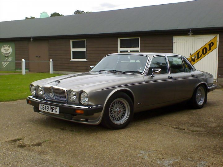 Pistonheads - The image shows a vintage Aston Martin DB4 front-driver, a timeless automobile with classic design features such as silver-gray paint, a grille radiator, and wire wheels. The car is parked outside a barn or garage, which has a green sign that reads "MACHINES & TOOLS" and a yellow sign with the word "LOP" on it. The sky appears overcast, suggesting a possibly cool or cloudy day. The setting is a rural, private driveway with a hint of a well-trimmed lawn.