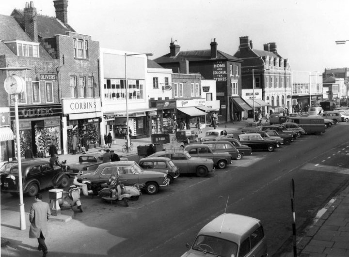 This car is driving me mad - Page 4 - Classic Cars and Yesterday's Heroes - PistonHeads UK - The image is a black and white photograph capturing a bustling city street scene. It's a busy urban area with numerous cars parked along the side of the road, and some in motion, creating a sense of movement and activity. The buildings lining the street appear to be shops or small businesses, suggesting a commercial district. Pedestrians are scattered throughout the image, adding to the overall hustle and bustle. The style of the vehicles and the clothing of the people suggest that this photo was taken in the mid-20th century.