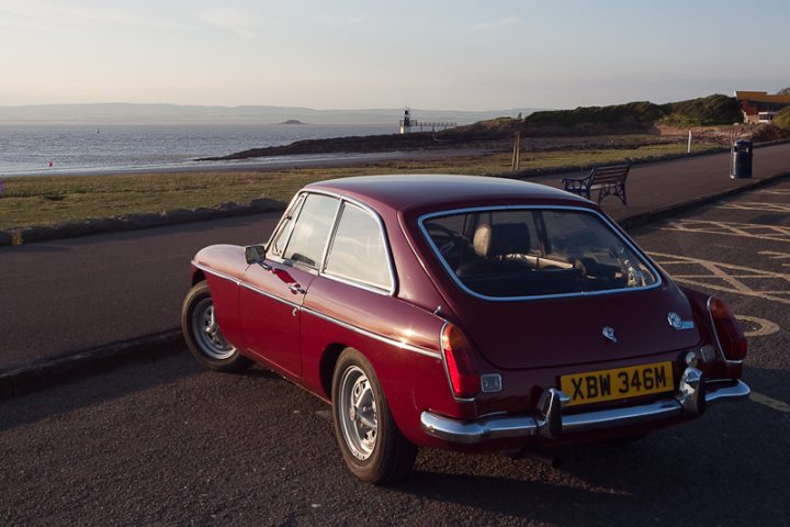 Who's got what in the South West - Page 1 - South West - PistonHeads - The image showcases a vintage car parked on the side of a road next to a body of water. The car is an older model, similar to those famous in the 1950s and 1960s, and is painted in a vibrant shade of red. The surroundings suggest a seaside location, with a bench visible on the adjacent path. The car's color contrasts beautifully with its natural environment. The setting appears serene and quiet, with no other vehicles or pedestrians in sight.