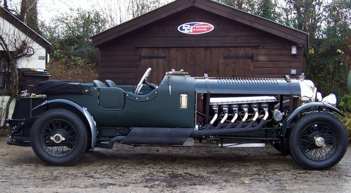 Handbuilt Bentley Specials by Vintage Racing - Page 2 - General Gassing - PistonHeads - The image features a vintage, dark green car with a large number of pipes on its neck, assuming it is a boat-like car. The car is a rarity, likely an antique, as indicated by the google map description. It is parked in front of a brown wooden garage, which is topped by a white roof. The setting suggests a private or rural area, with no other vehicles or buildings immediately surrounding the garage. The car itself has a weathered appearance, adding to its charming vintage vibe.