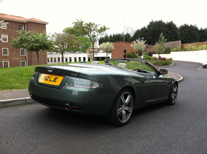 Clear rear lights? - Page 1 - Aston Martin - PistonHeads - The image captures a sleek, black Aston Martin car parked on the right side of a gray asphalt road. The license plate of the car reads "NOJIE." The car is positioned in front of a manicured lawn, with a clear view of a brick building to the left and lush green trees in the background. The scene suggests a suburban or urban setting during the daytime due to the natural light present. The car's shiny surface and sporty design indicate it is well-maintained and likely a luxury model. The overall atmosphere is calm and orderly.