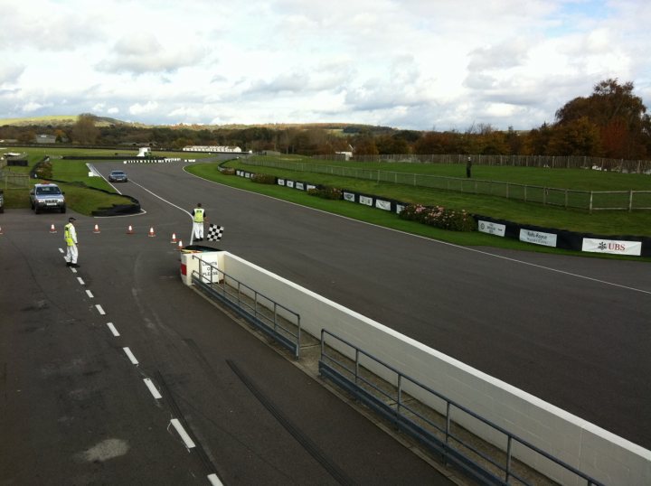 Goodwood Aston day - Page 1 - Aston Martin - PistonHeads - The image shows a temporary checkpoint on a road, possibly during a racing event or a special traffic situation requiring lane closures. A few traffic cones have been set up with white and black stripes, and there are two uniformed individuals lowering a barricade to perform some sort of duty. The road is wide and appears to be at a crossroads, with a track or stadium directly ahead of the checkpoint. Fitted gates are seen, indicating controlled entry or exit on this road. The backdrop features a lap of a golf course, suggesting that the location might be near a resort or recreational area. The sky is overcast with thick clouds indicating a possibility of rain, as there may be flagsticks suggestive of racing conditions.