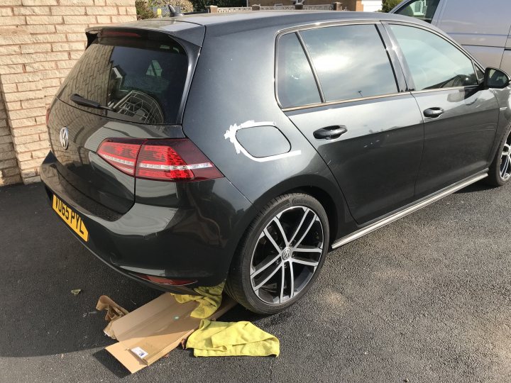 Golf MK7 petrol cap lettering stuck shut - Page 1 - Audi, VW, Seat & Skoda - PistonHeads - The image shows a dark-colored SUV parked outdoors. Its bodywork is freshly repaired, as evidenced by the visible scratches that have been sanded down and primed. A pair of gloves can be seen on the ground near the car's front wheel. The vehicle appears to be in a residential area, as indicated by the presence of a brick building and other houses in the background. There is no text visible in the image.