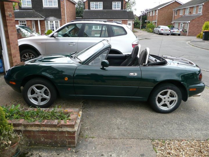 Pistonheads Grounds Hunting - The image features a vibrant green, two-door convertible sports car parked on a driveway. The car has a black soft top roof and the wheels are silver alloys. To the left of the car, there is a medium-sized white SUV parked. A row of houses with similar roof tiles forms a backdrop to the scene. The image captures a typical day with clear visibility and the photograph has a casual, real-life setting.