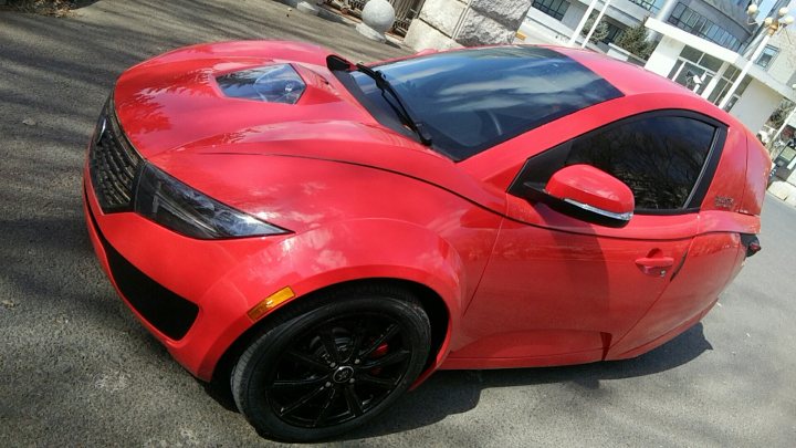 Pistonheads - The image shows a vibrant red sports car parked on a city street. The vehicle has a sleek, aerodynamic design with prominent wheel arches and a low, wide stance, indicative of a high-performance model. It features tinted windows, a spoiler on the rear decklid, and large, multi-spoke alloy wheels. There are visible custom modifications such as additional body parts that suggest this car has been significantly altered for enhanced aesthetics or performance capabilities. The car is parked next to a building with a glass front, under what appears to be a bright, sunny sky.