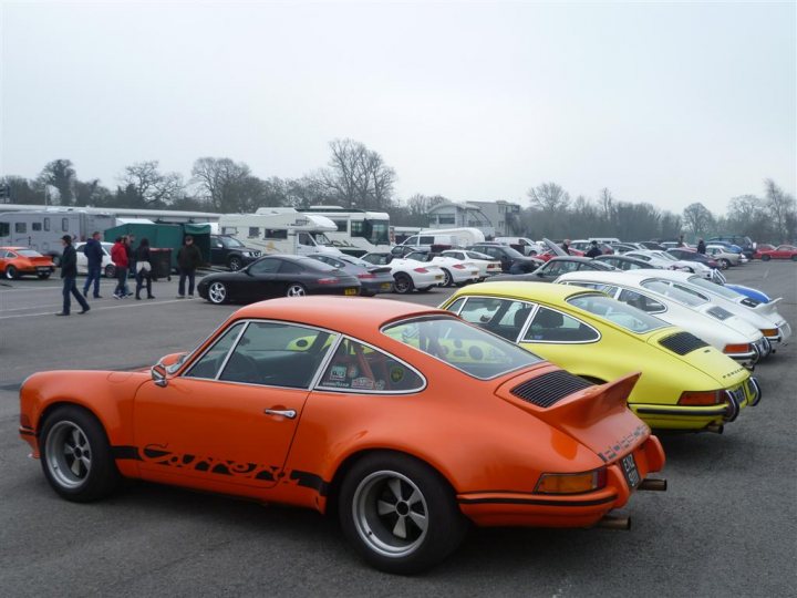 14th Annual Oulton Park RS day - Page 6 - Porsche General - PistonHeads - The image depicts a vibrant scene of a car show, where a row of colorful sports cars is on display. The cars, painted in shades of orange, yellow, and white, are parked side by side, their sleek designs and vivid hues drawing the eye. Each car is unique, exhibiting various makes and models, including some vintage models. A group of people are present, walking around and taking in the spectacle, lending a sense of scale to the image, highlighting the size and stature of these impressive vehicles. The perspective of the photo is from a slightly elevated angle, looking down on the cars and the crowd, allowing for a comprehensive view of the scene.