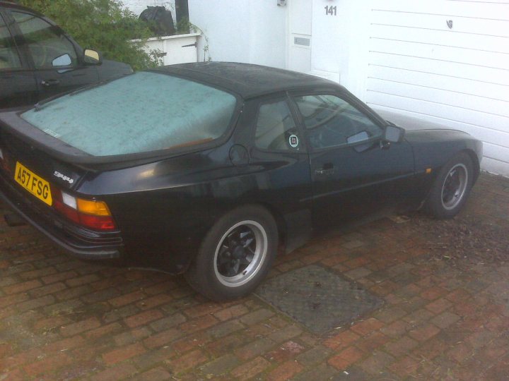 Love Pistonheads Hate - This image shows a dark-colored sports car parked on a red brick driveway. The car features a glass rear window (sometimes referred to as a backlight), a targa or dual cockpit top if any other make, a rear mudflap, a limited slip differential (LSD), and front and rear aerodynamic splitters. The asymmetrical exhaust features a dual pipe with a single pipe cutout on the right side, and there are some small markings or decals visible on the hood. The background suggests a residential area with a white house and a car reflected in its windows.