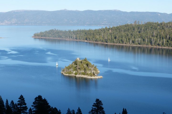 A train traveling over a bridge over a river - Pistonheads - The image presents a serene lake landscape. Dominating the scene is a small island, densely covered in lush trees, with a few bare patches providing a contrast to the greenery. The island appears to be of volcanic origin. The lake is bordered by a backdrop of untouched trees on the left, providing a sense of seclusion. In the distance, dotting the horizon, are a few boats, navigating the tranquil waters. The sky above is a clear blue with a few clouds scattered in the distance. The overall scene is one of peace and natural beauty.